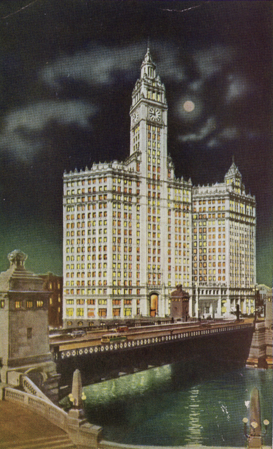 A color image of the Wrigley Building at night. The stark white building rises up from behind a bridge over a river. All of its windows are illuminated from the interior and glow orange or yellow. The upper portions of the building have classical details. In the center is a large square tower with a clock. A white full moon shines through the clouds.